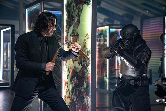 A man in a black suit uses a wooden tool to fight an armored figure in a dark, modern room.