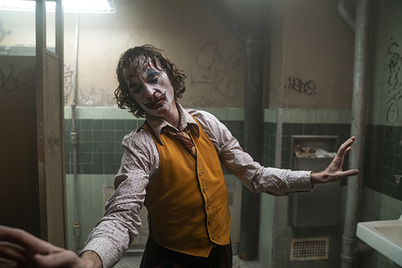 A man with clown makeup and a yellow vest dances in a grimy, tiled bathroom.