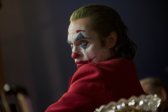 A man with white face paint, blue diamonds over his eyes, and a red nose looks back over his shoulder.