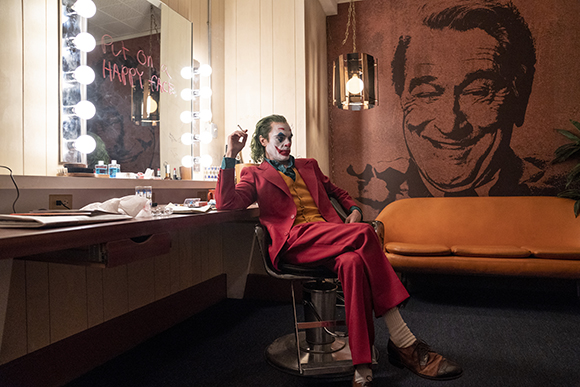 A man in a red suit and clown makeup sits in a chair in a dressing room with a large portrait on the wall.