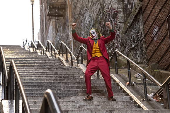 A man in clown makeup and a red suit dances with arms raised on a long outdoor concrete staircase.
