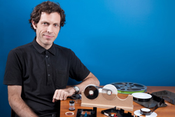A man in a black shirt sits at a table with film reels and equipment in front of a blue background.
