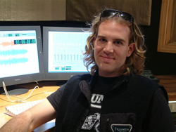 A man with wavy hair and sunglasses on his head sits before two computer monitors in a studio.