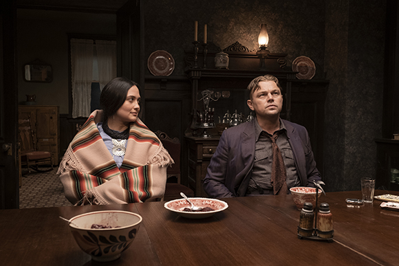 A woman in a patterned blanket and a man in a dark suit sit at a dining table in a dimly lit room.
