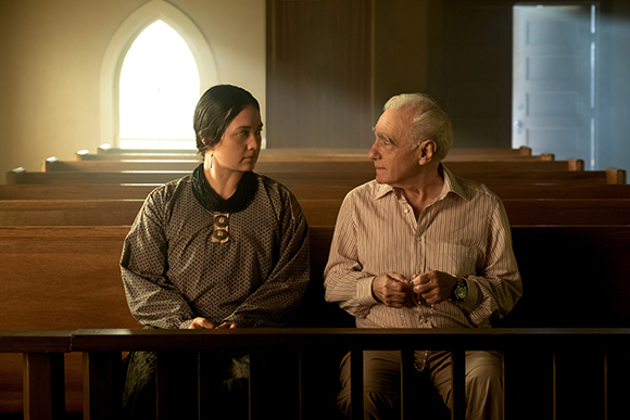 A young woman in dark clothing and an older man in a striped shirt sit together in a church pew.