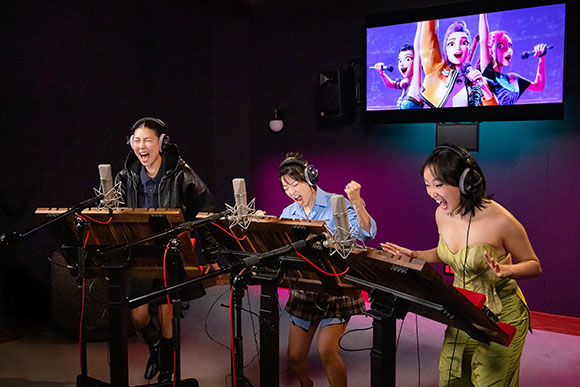Three women sing and play keyboards in a dark studio while watching an animated music video on a screen.