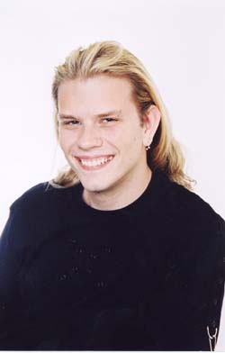 A smiling young man with long blonde hair wearing a black shirt against a white background.