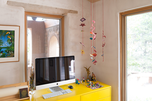 A computer monitor sits on a bright yellow desk next to a window with hanging pink decorations.