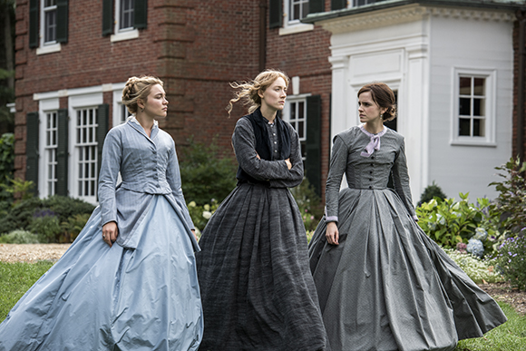 Three women in long, voluminous 19th-century dresses stand in a garden in front of a brick house.