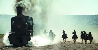 A black steam train emitting white smoke as several people on horseback ride alongside it on a dirt path.