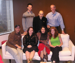 Six diverse people posing together, some standing and some sitting on a white sofa.