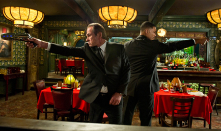 Two men in dark suits aim handguns in opposite directions inside a dimly lit restaurant.