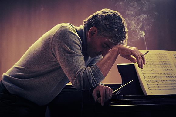 A man rests his head on his arms atop a piano with sheet music visible.