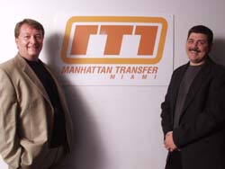 Two men in blazers smiling in front of a Manhattan Transfer Miami logo.
