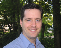 A man with dark hair and a blue shirt smiling outdoors with green trees in the background.
