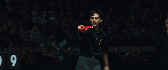 A man in a dark polo shirt swings a red table tennis paddle in a dimly lit arena.