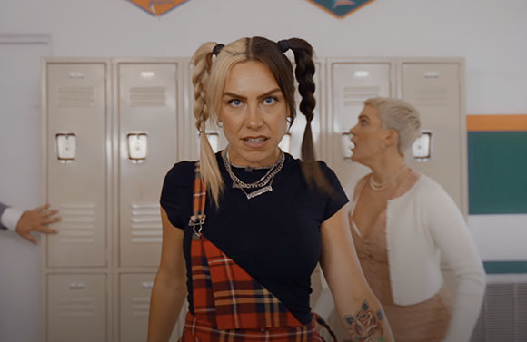 A woman with blonde and brown pigtails wears a black shirt and plaid overalls in a school hallway.