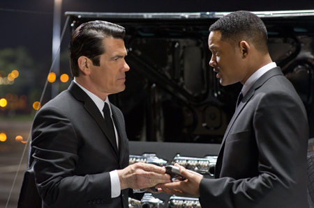 Two men in black suits face each other while holding a small object near an open car trunk at night.