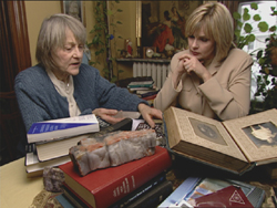 An older woman in a blue sweater shows an open book to a blonde woman in a tan blazer.