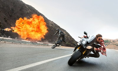 Two motorcyclists speed along a winding mountain road as a large fireball erupts in the background.