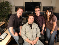 Four people smiling and posing in a professional recording studio with monitors and equipment.