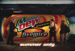 A giant Mountain Dew LiveWire can sits in a dark space with three people walking past it.