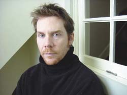 A man with light brown hair and a reddish beard wearing a black turtleneck leans against a white window frame.