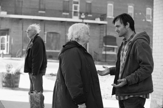 A young man and an elderly woman stand facing each other in conversation outdoors.