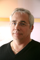 Close-up portrait of a middle-aged man with short gray hair, wearing glasses and a black V-neck shirt with a gold chain.