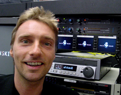 A man smiling in front of a rack of professional audio mixing and playback equipment.