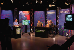 People sit at desks in a television studio with large screens and bright lighting.