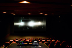 A dark movie theater showing a bright scene on a large screen above rows of red seats.