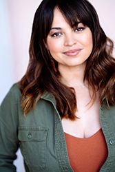 A woman with wavy dark hair and bangs wearing an olive green shirt over a rust-colored top.