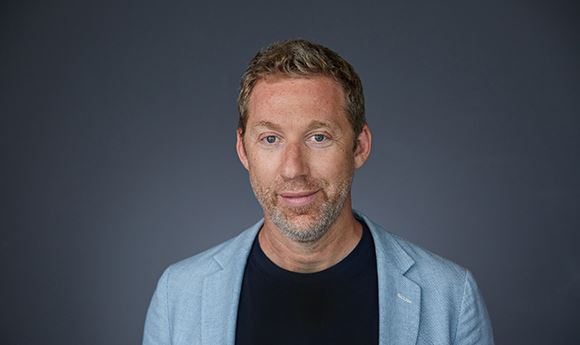 A man with short blond hair and light stubble wearing a black shirt and light blue blazer against a dark background.