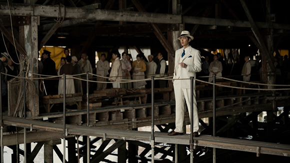 A man in a white suit and straw hat stands on a wooden pier with a crowd of people in the background.