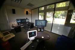 Wide-angle view of an office with several computer monitors, desks, and large windows overlooking trees.