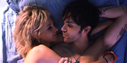 A blonde woman and a dark-haired man lie close together on blue bedding in dim lighting.