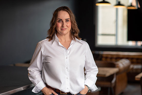 A woman with wavy brown hair wearing a white long-sleeved button-down shirt, standing in a dimly lit restaurant.