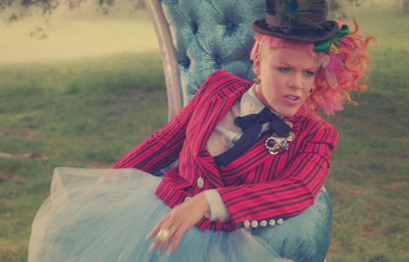 A person with pink hair and a top hat wearing a red striped jacket and blue tulle skirt sits in a blue chair.
