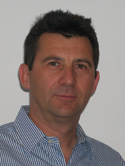 A man with dark hair wearing a blue and white striped button-down shirt against a plain background.