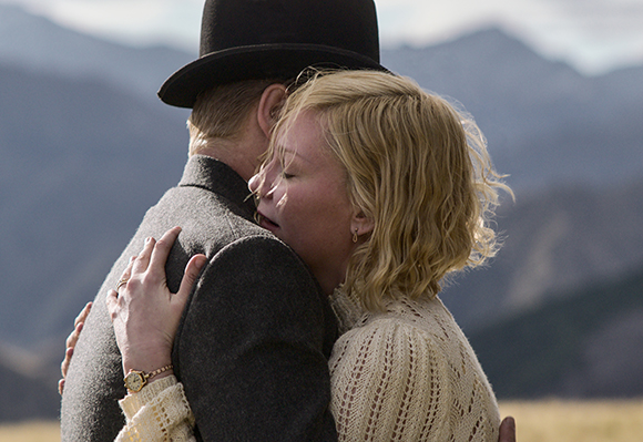 A man in a black top hat and grey coat hugs a blonde woman in a white lace dress against a mountain backdrop.