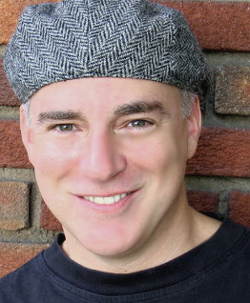 A smiling man in a grey herringbone flat cap against a brick wall.