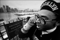 A man in glasses and a Brooklyn cap talks on a mobile phone by a waterfront city skyline.