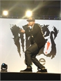 A man in a dark jacket and cap performs on a stage in front of a large graphic backdrop.