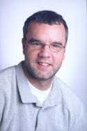 A man with short dark hair and glasses smiling against a light background.