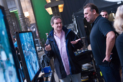 A man in a dark jacket gestures with both hands while talking to a man in a black polo shirt on a film set.