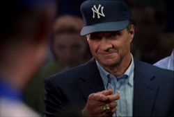 A man wearing a navy Yankees cap and suit jacket points his finger forward.