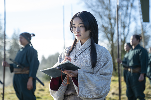 A woman in a patterned kimono holds an open notebook while standing outdoors near several men with spears.