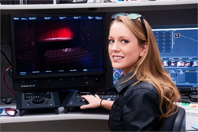 A smiling woman sits at a desk with large monitors displaying video footage and editing software.