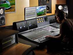 A person sits at a large mixing console with dual monitors and studio speakers in a recording studio.